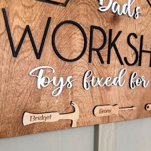 May include: Wooden sign with the text "Dad's Workshop Toys fixed for" in white lettering. The sign also features a hammer and screwdriver with the names "Bridget" and "Bronx" engraved on them.