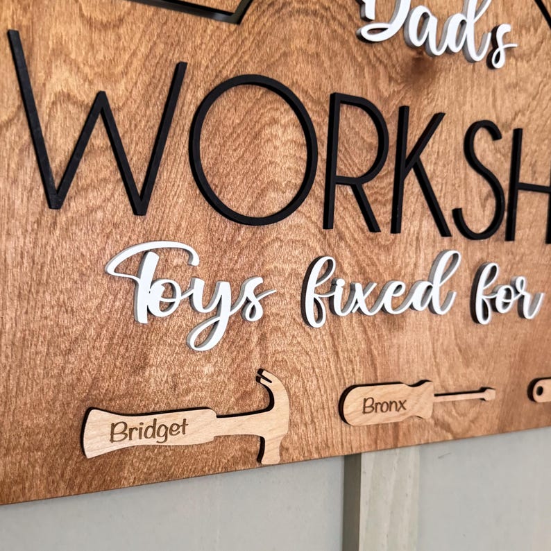 May include: Wooden sign with the text "Dad's Workshop Toys fixed for" in white lettering. The sign also features a hammer and screwdriver with the names "Bridget" and "Bronx" engraved on them.