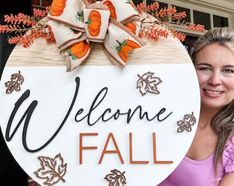 Puede incluir: Un letrero redondo de madera blanca con las palabras "Welcome Fall" en letras negras y naranjas. El letrero está decorado con una cinta de otoño marrón y naranja con calabazas y hojas. El letrero lo sostiene una persona que lleva una camisa morada.