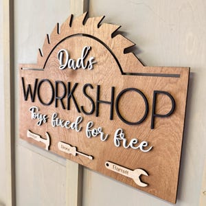May include: A wooden sign with the text "Dad's Workshop Toys fixed for free" in white lettering with black outlines. The sign has a saw blade shape at the top and includes three tools with names "Jordan", "Bronx", and "Barrett" written on them.