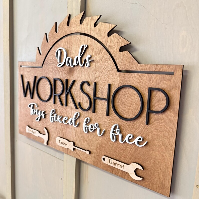 May include: A wooden sign with the text "Dad's Workshop Toys fixed for free" in white lettering with black outlines. The sign has a saw blade shape at the top and includes three tools with names "Jordan", "Bronx", and "Barrett" written on them.