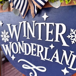 May include: A round wooden sign with the words "WINTER WONDERLAND" in white, with snowflake and star accents. The sign is navy blue with a decorative bow at the top. The bow has a striped pattern and a burlap texture.