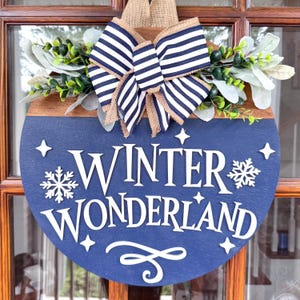 May include: A round wooden sign with a navy blue base and the words "WINTER WONDERLAND" in white. The sign is decorated with a striped bow, greenery, and a burlap ribbon. The sign is designed for winter or holiday decor.