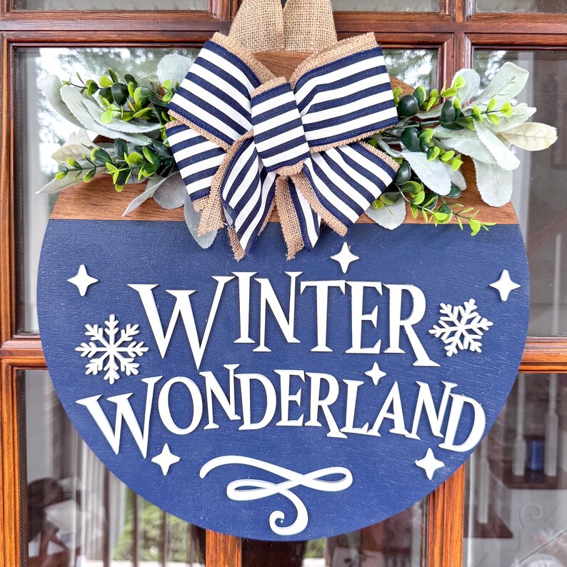 May include: A round wooden sign with a navy blue base and the words "WINTER WONDERLAND" in white. The sign is decorated with a striped bow, greenery, and a burlap ribbon. The sign is designed for winter or holiday decor.
