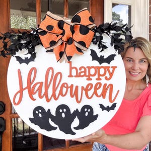 May include: A circular white Halloween sign with orange lettering reading "Happy Halloween." The sign features black bats, ghosts, and an orange and black polka dot bow. Black leaves are attached to the top of the sign.