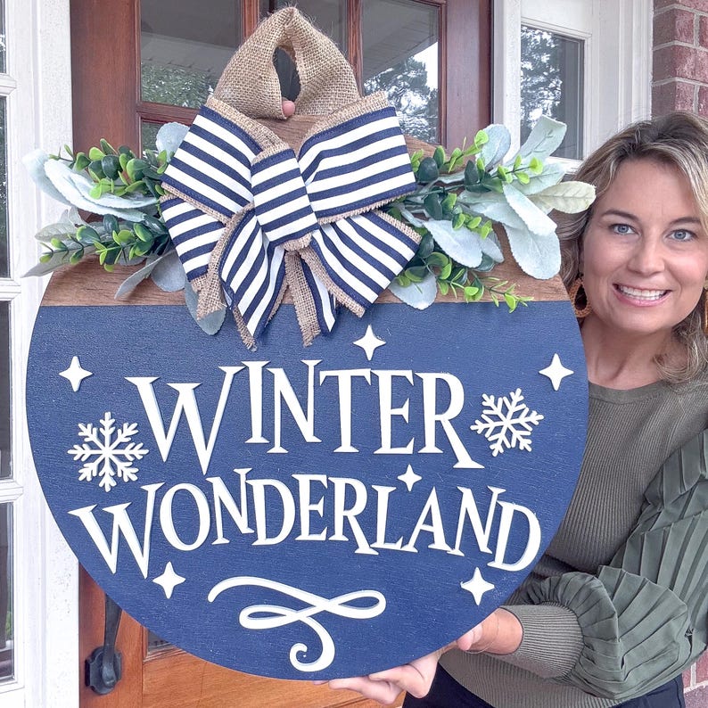 May include: A round wooden sign with a navy blue painted bottom half and a natural wood top half. The sign reads "WINTER WONDERLAND" in white script and features a snowflake and star accents. A burlap and striped bow and greenery complete the festive design.