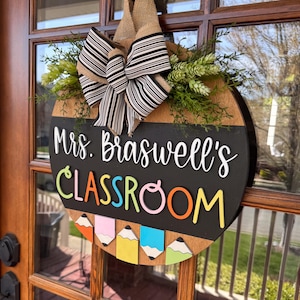Op de afbeelding: Een ronde houten deurhanger met een krijtbord. Het krijtbord zegt "Mrs. Braswell's Classroom" in witte letters. De bovenkant van de deurhanger is versierd met een zwart-wit gestreepte strik en groen blad.