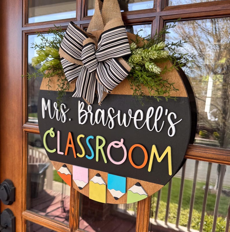 Op de afbeelding: Een ronde houten deurhanger met een krijtbord. Het krijtbord zegt "Mrs. Braswell's Classroom" in witte letters. De bovenkant van de deurhanger is versierd met een zwart-wit gestreepte strik en groen blad.