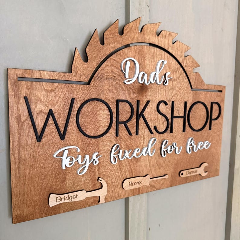 May include: A wooden sign with a saw blade design, reading "Dad's Workshop Toys fixed for free".  The sign has three tool-shaped hooks with the names "Bridget", "Bronx", and "Barnett" written on them.