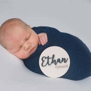 K&ouml;nnte beinhalten: Ein neugeborenes Baby liegt in einer blauen Decke gewickelt und schl&auml;ft. Ein rundes, wei&szlig;es Holzschild mit dem Namen "Ethan Edward" liegt neben dem Baby.