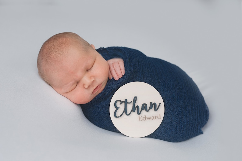 K&ouml;nnte beinhalten: Ein neugeborenes Baby liegt in einer blauen Decke gewickelt und schl&auml;ft. Ein rundes, wei&szlig;es Holzschild mit dem Namen "Ethan Edward" liegt neben dem Baby.
