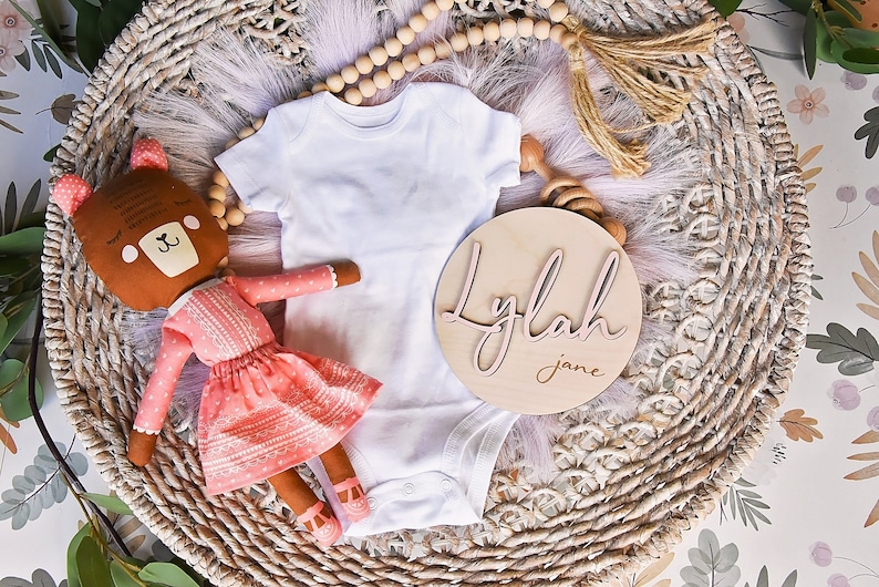 K&ouml;nnte beinhalten: Ein wei&szlig;er Strampler, eine braune und rosafarbene Puppe und ein Holzschild mit dem Namen "Lylah Jane" darauf. Die Gegenst&auml;nde sind in einem geflochtenen Korb mit einer rosa-wei&szlig;en, flauschigen Textur angeordnet.