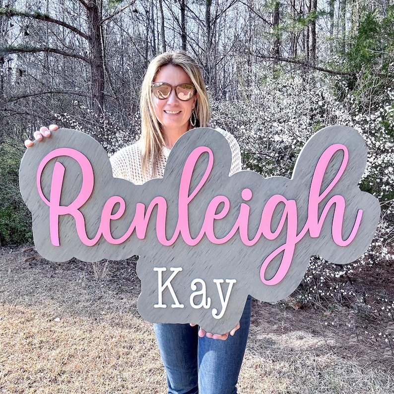 May include: A gray wooden sign with the name "Renleigh Kay" written in pink cursive letters.