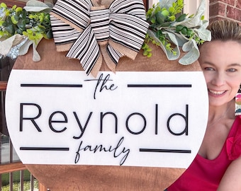 Puede incluir: Un colgador de puerta redondo de madera con fondo blanco y letras negras que dice "The Reynold Family". Un lazo a rayas blanco y negro con un centro de arpillera está unido a la parte superior del colgador.