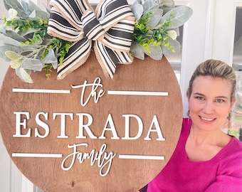 Puede incluir: Un letrero redondo de madera con el texto "The Estrada Family" en letras blancas. El letrero está decorado con un lazo a rayas blanco y negro y vegetación.