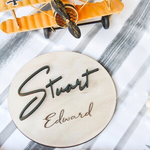 K&ouml;nnte beinhalten: Ein rundes Holzschild mit den Namen "Stuart Edward" in schwarzer Schrift auf hellem Holzgrund.