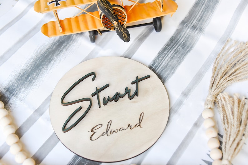 K&ouml;nnte beinhalten: Ein rundes Holzschild mit den Namen "Stuart Edward" in schwarzer Schrift auf hellem Holzgrund.