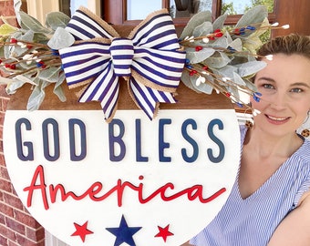 Può includere: Un cartello rotondo in legno bianco e marrone con il testo "God Bless America" in lettere blu e rosse. Il cartello è decorato con un fiocco blu e bianco a righe e un rametto di bacche bianche e rosse.