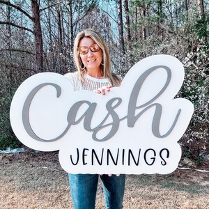 May include: A white sign with the name "Cash Jennings" written in gray and black lettering. A person is holding the sign in front of a green background.