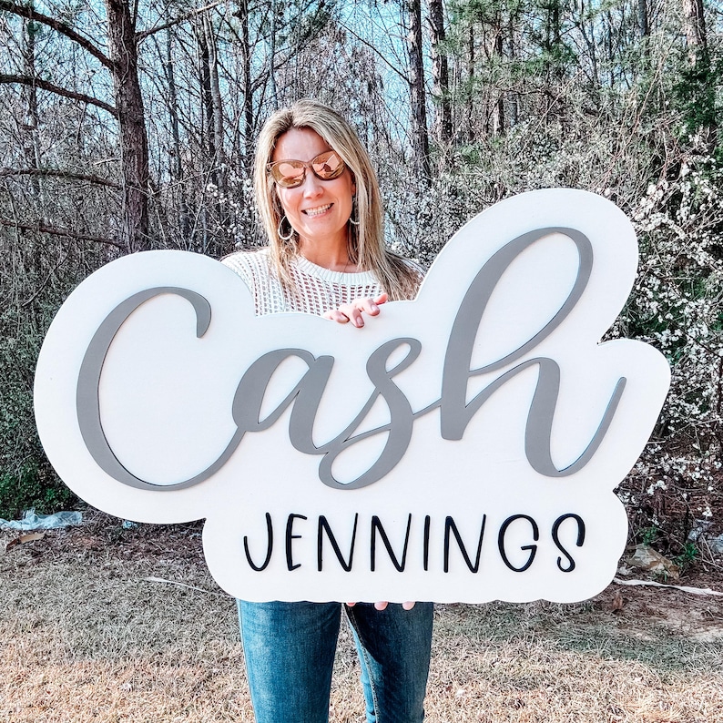 May include: A white sign with the name "Cash Jennings" written in gray and black lettering. A person is holding the sign in front of a green background.