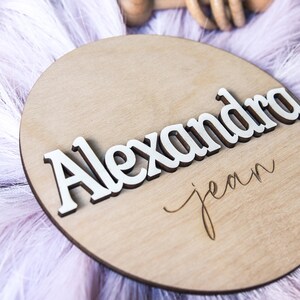 K&ouml;nnte beinhalten: Ein rundes Holzschild mit dem Namen "Alexandra Jean" in wei&szlig;en Buchstaben. Das Schild ist mit einem wei&szlig;en, flauschigen Hintergrund verziert.