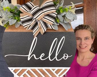 Puede incluir: Cartel de puerta redondo de madera con superficie pintada en negro y marrón. La palabra "hello" está escrita en blanco. Un lazo decorativo con rayas blancas y negras y cinta de arpillera está en la parte superior, junto con vegetación artificial. La parte inferior tiene un diseño geométrico.