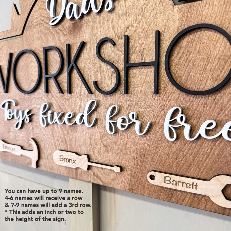 May include: A wooden sign with the text "Dad's Workshop Toys fixed for free" in white lettering. The sign has a brown wood grain finish and features a variety of tools, including a hammer, screwdriver, and wrench. The sign is personalized with the names "Bridget", "Bronx", and "Barrett".