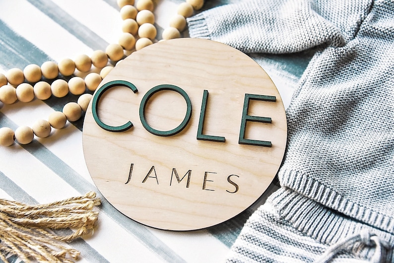 K&ouml;nnte beinhalten: Ein rundes Holzschild mit dem Namen "COLE JAMES" in gr&uuml;nen Buchstaben. Das Schild befindet sich auf einem gestreiften Hintergrund mit einer Schnur aus Holzperlen und einem St&uuml;ck Jute.