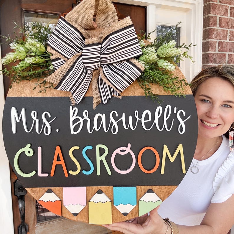 Op de afbeelding: Een ronde houten deurhanger met een krijtbord. Het krijtbord zegt "Mrs. Braswell's Classroom" in witte letters. De letters in het woord "Classroom" zijn omrand met verschillende kleuren. Er hangen vijf kleurpotloden aan de onderkant van de deurhanger.