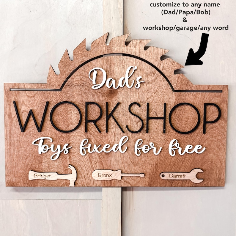 May include: Wooden sign with a saw blade design, reading "Dad's Workshop Toys fixed for free". The sign has three smaller wooden pieces with the names "Bridget", "Bronx", and "Barrett" and images of a hammer, screwdriver, and wrench.