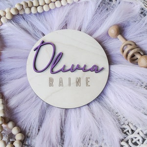 K&ouml;nnte beinhalten: Ein Holz-Kreis mit dem Namen "Olivia Raine" in lila Schrift. Der Kreis ist von einem flauschigen, helllila Material und einem Holzrassel umgeben.