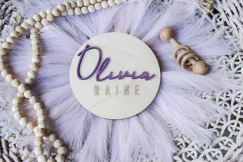 K&ouml;nnte beinhalten: Ein Holz-Kreis mit dem Namen "Olivia Raine" in lila Schrift. Der Kreis ist von einem flauschigen, helllila Material und einem Holzrassel umgeben.