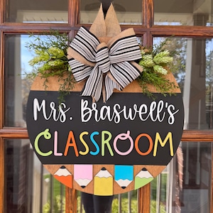Op de afbeelding: Een ronde houten deurhanger met een schoolbord oppervlak. Het schoolbord zegt "Mrs. Braswell's Classroom" in witte letters. De deurhanger is versierd met een zwart-wit gestreepte strik, groen blad en kleurrijke potloden.
