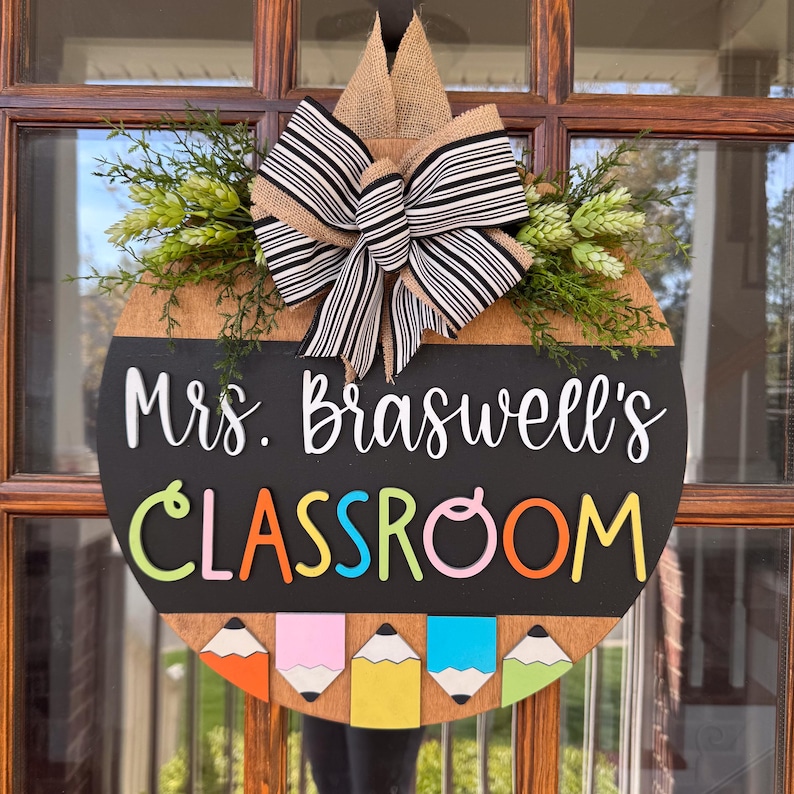 Op de afbeelding: Een ronde houten deurhanger met een schoolbord oppervlak. Het schoolbord zegt "Mrs. Braswell's Classroom" in witte letters. De deurhanger is versierd met een zwart-wit gestreepte strik, groen blad en kleurrijke potloden.