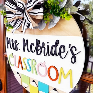 Op de afbeelding: Een rond, wit houten bord met de tekst "Mrs. McBride's Classroom" in zwarte letters. Het bord is versierd met een zwart-wit gestreepte strik, groene bladeren en kleurpotloden.