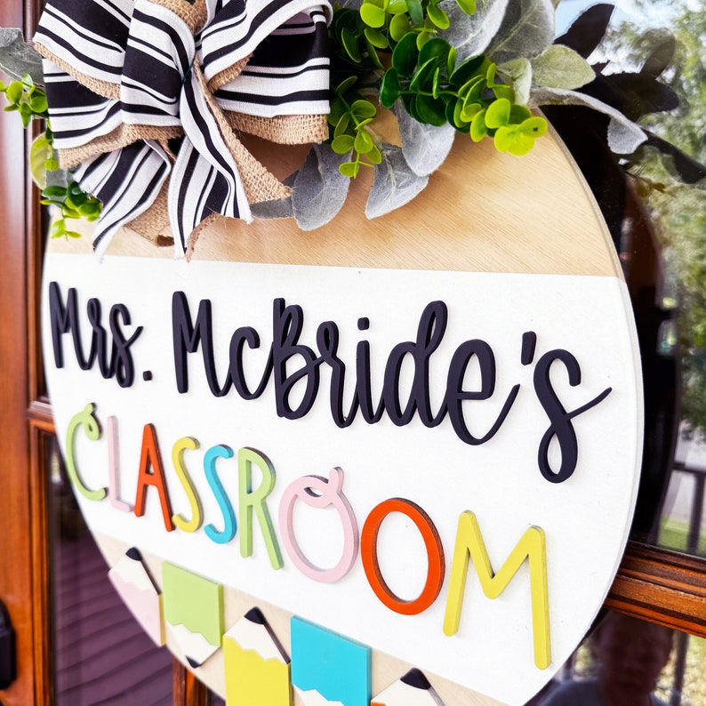 Op de afbeelding: Een rond, wit houten bord met de tekst "Mrs. McBride's Classroom" in zwarte letters. Het bord is versierd met een zwart-wit gestreepte strik, groene bladeren en kleurpotloden.