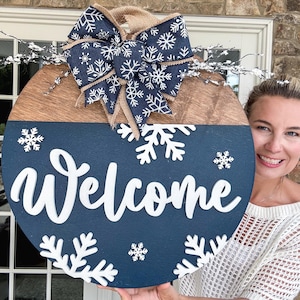 May include: A round wooden door hanger with a blue and brown woodgrain design. The top half is brown woodgrain and the bottom half is painted blue. The word "Welcome" is written in white cursive letters on the blue background. The door hanger is decorated with white snowflakes and a white and blue bow with a burlap center.