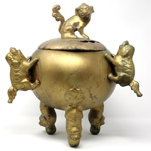 May include: A gold-colored incense burner with a round body, three legs, and a lid. The lid is topped with a small monkey figurine. Two other monkey-like figures are attached to the sides.