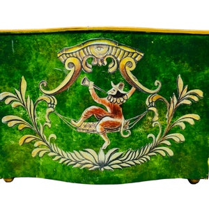 May include: A green painted box with a decorative design. The box features a central illustration of a figure playing a horn, surrounded by ornate gold and white floral accents. The box has gold trim and small feet.