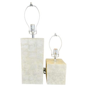 May include: Two decorative table lamps. The lamp bases are rectangular and feature a textured, off-white shell-like design. Each lamp has a silver-colored neck and a silver-colored lamp holder. The lamps are of different sizes.