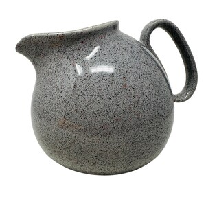 May include: A speckled gray ceramic pitcher with a rounded body and a curved handle. The pitcher has a spout for pouring and is covered in small black and red flecks. The pitcher is a kitchenware item.