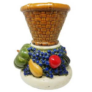 May include: A ceramic basket-shaped container with a woven texture, brown glaze, and a base decorated with grapes, a red apple, and other fruits. The base is off-white with green accents.