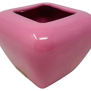 May include: A glossy, pink ceramic vase with a square opening and rounded sides. The vase has a smooth, reflective surface and a small, circular sticker on the side. The interior of the vase is a darker shade of pink.