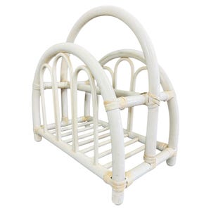 May include: Off-white rattan magazine rack with a curved handle and arched supports. The rack has a rectangular base with horizontal slats and vertical dividers. The overall design is a vintage style.