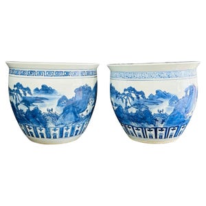 May include: Two matching white ceramic planters with a blue landscape design. The planters feature a wide, rounded shape with a detailed blue and white painted scene of mountains, trees, and bridges. The rims and bases have decorative patterns.