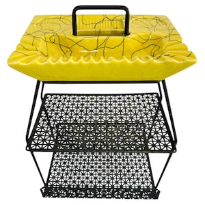 May include: A vintage yellow ashtray with a black handle and abstract black lines, set on a black metal stand with two mesh shelves. The ashtray has a wavy edge.