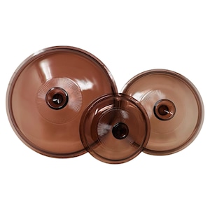 May include: Three brown glass lids of varying sizes are arranged against a white background. Each lid has a central knob and a radial pattern emanating from the center. The lids appear to be for cookware.