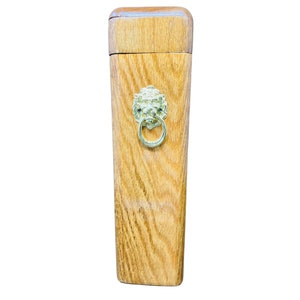 May include: A wooden box with a lid, featuring a light brown finish and a decorative brass lion's head with a ring handle. The box has a rectangular shape and a slightly tapered design.