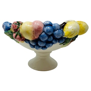 May include: A white ceramic pedestal fruit bowl with a colorful assortment of sculpted fruit. The bowl features grapes, peaches, lemons, and berries. The fruit is painted in realistic colors and sits atop a white pedestal base.