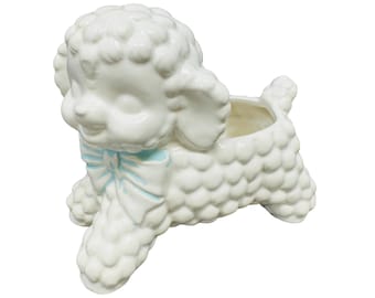 May include: A white ceramic poodle planter with a light blue bow. The poodle has a textured, curly coat and a smiling face. The planter is designed to hold small plants or flowers.